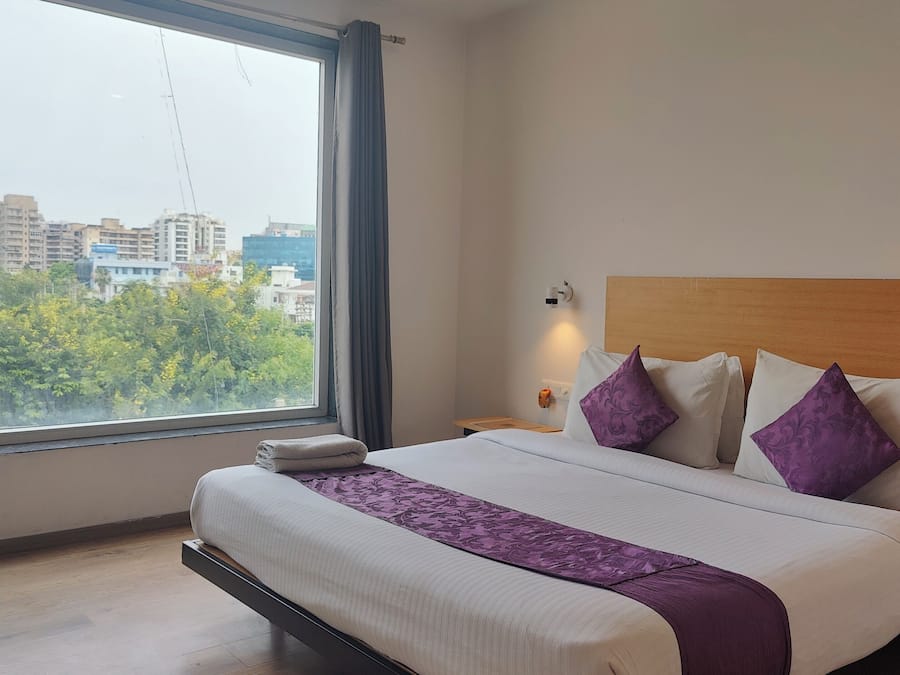 A hotel room with a large window, a bed with white bedding and purple pillows, a wooden headboard, and a nightstand with a lamp.