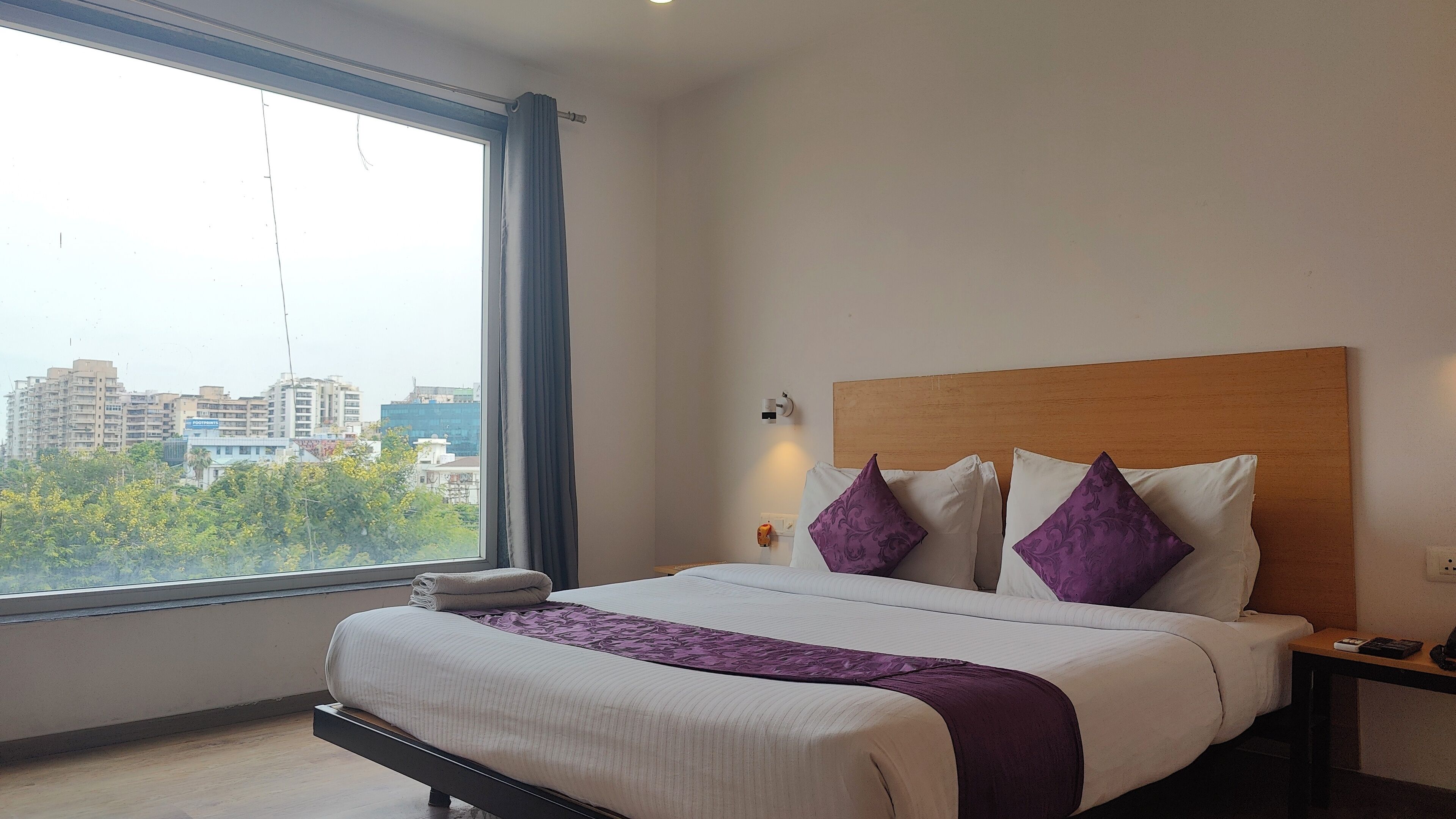 Una habitación de hotel con un ventanal grande, una cama con ropa de cama blanca y morada, un cabecero de madera y una mesita de noche con una lámpara.