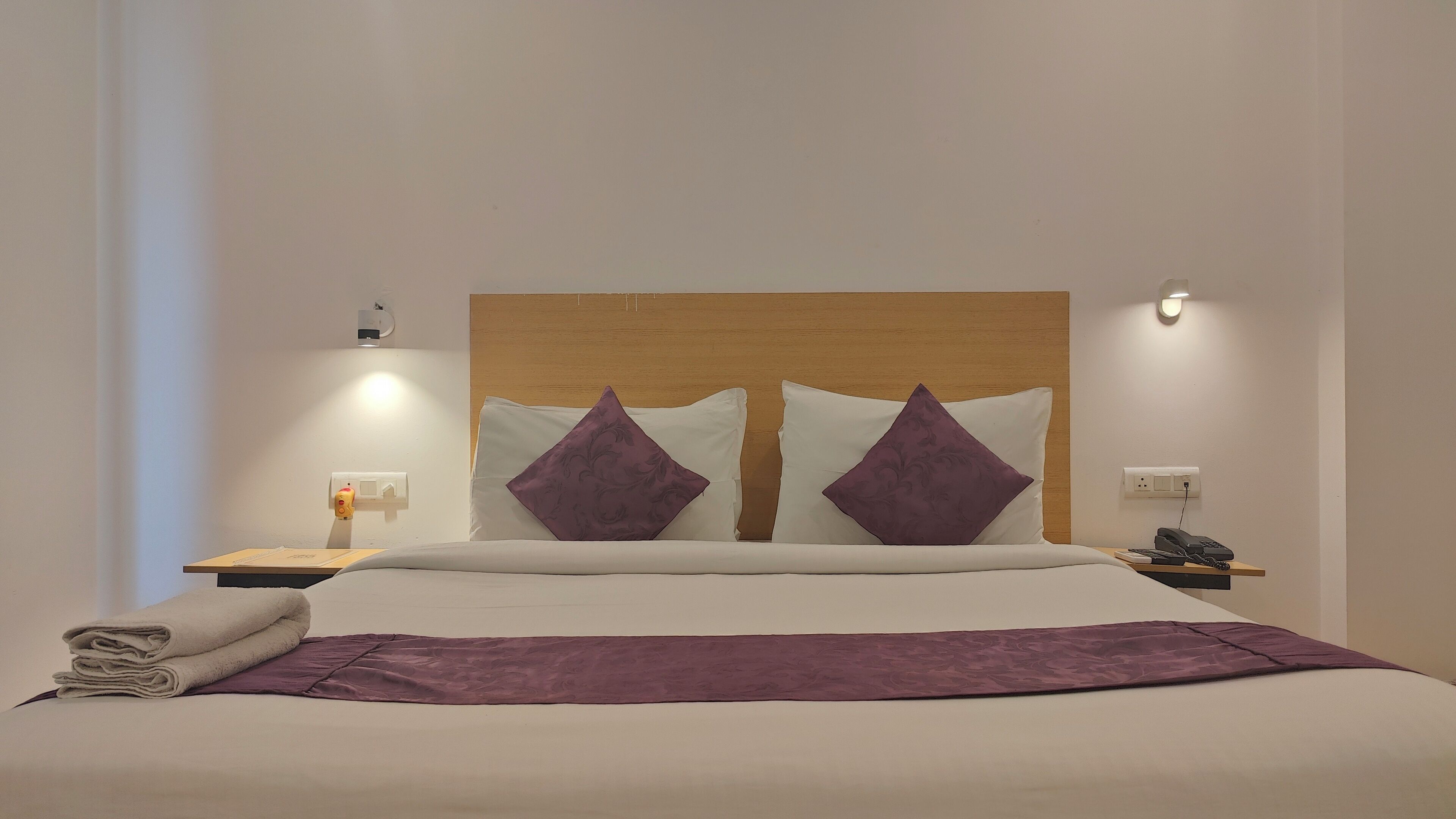 A neatly made bed with a purple throw blanket, two purple pillows, and a wooden headboard. There is a bedside table with a lamp, a phone, and a small plant.