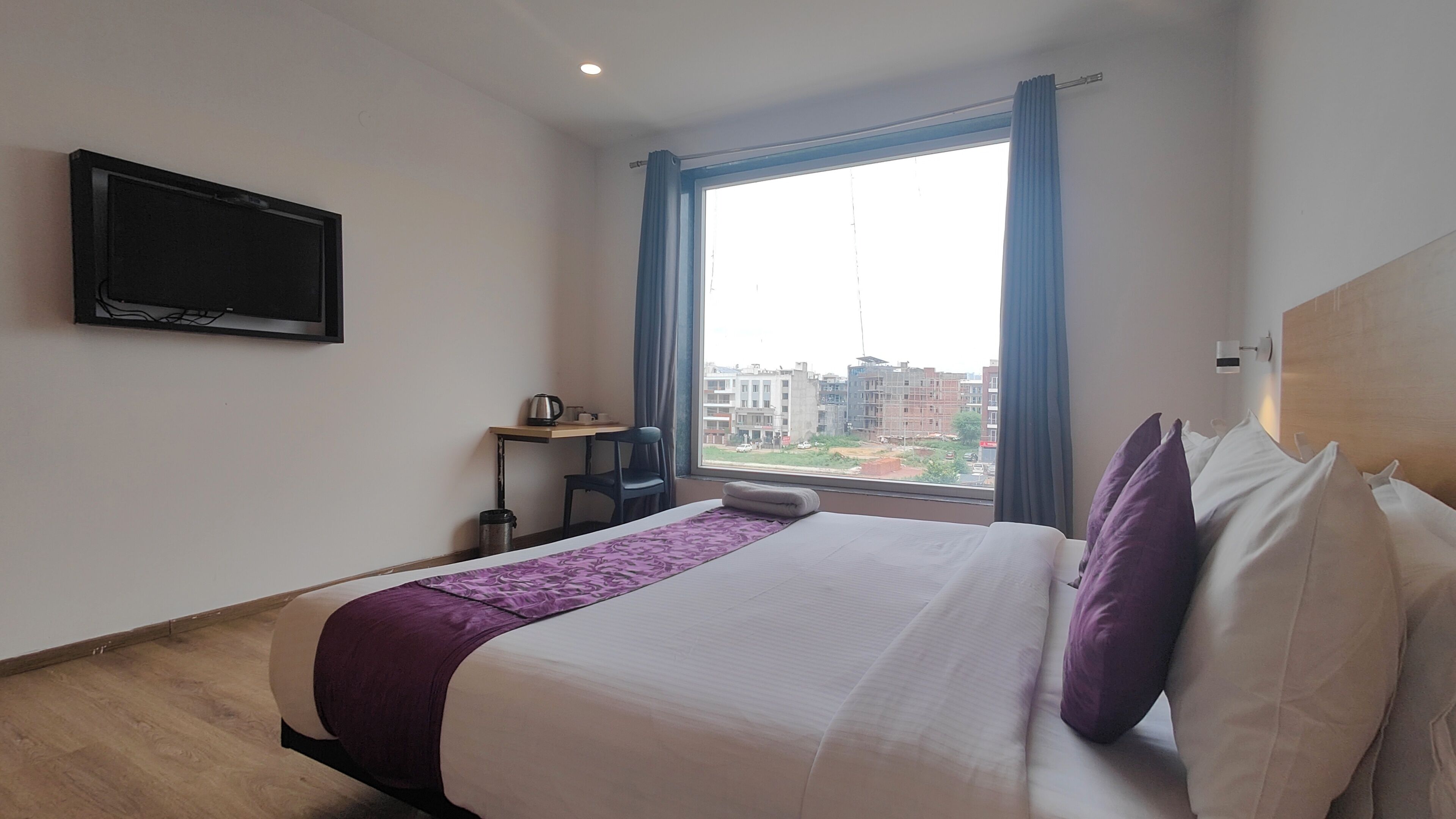 A hotel room with a bed, a desk, a chair, a television mounted on the wall, and a window with a view of buildings.