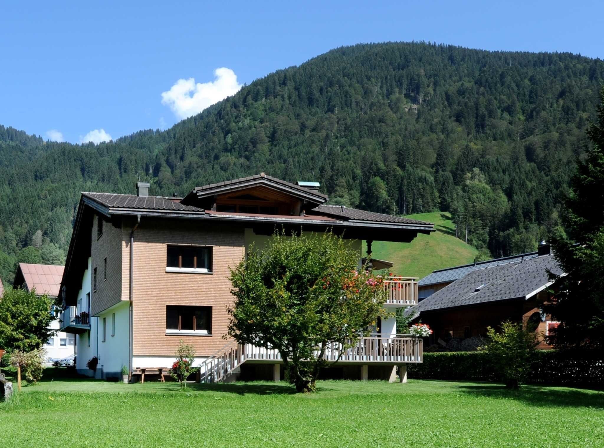 A two-story building with a balcony, surrounded by a grassy yard and a forested hill.