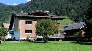 Un bâtiment à deux étages doté d’un balcon, entouré d’une cour verdoyante et d’une colline boisée.