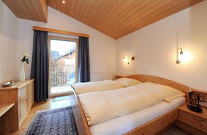 A room with two beds, a wooden ceiling, a window with curtains, a bedside table, and a nightstand.