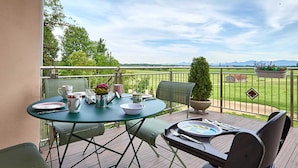 Een glazen tafel gedekt met borden, bekers en een karaf op een balkon met uitzicht op een groen landschap en bergen.