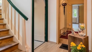 Een trap met houten treden en groene leuningen, leidend naar een kamer met een houten vloer, een zwarte deurmat en een potplant met gele bloemen.