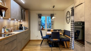 Een moderne keuken met houten kastjes, een zithoek met een blauwe bank en stoelen, een klok aan de muur en een raam met gordijnen.