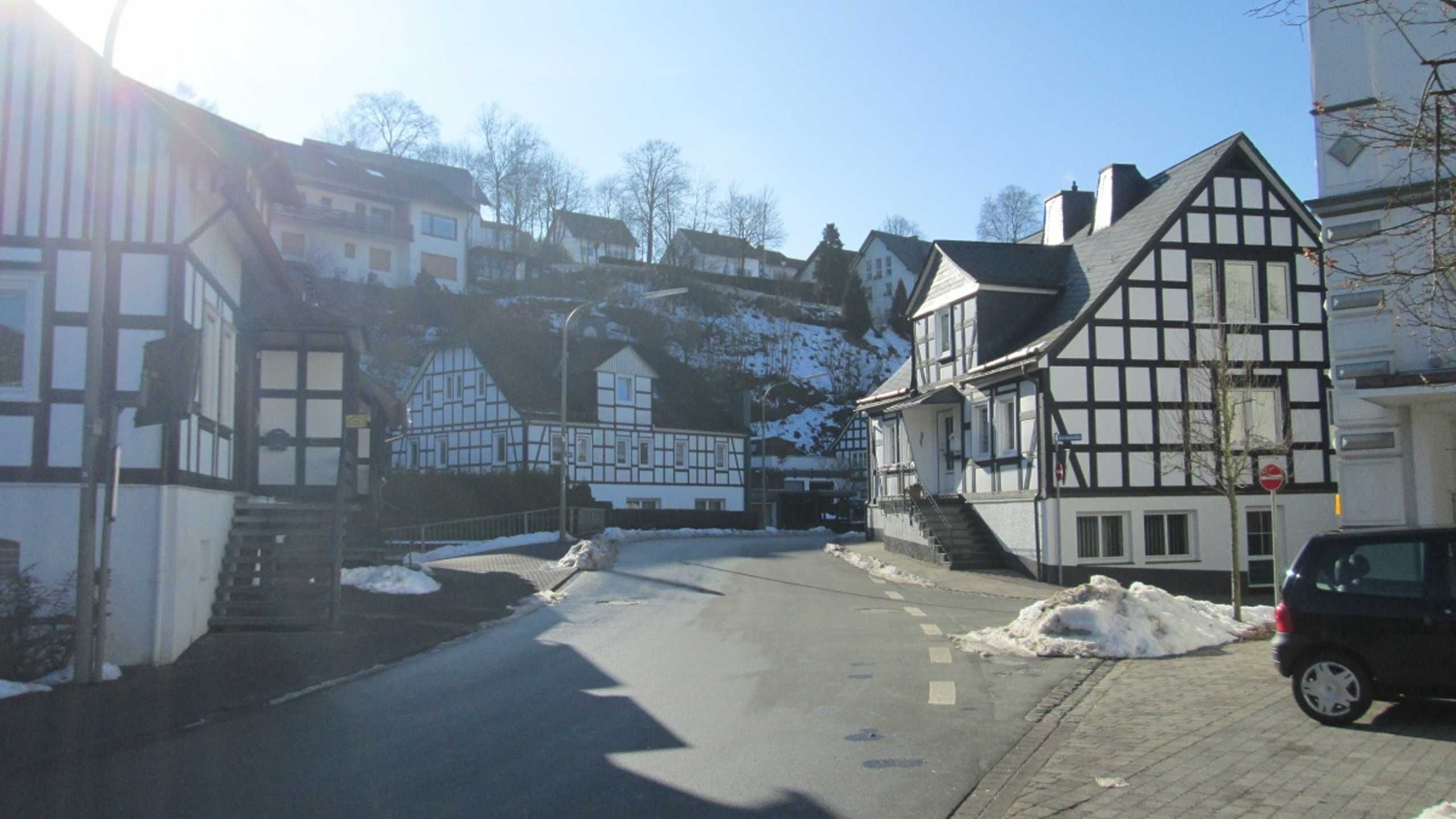 A street with traditional half-timbered houses, a car parked on the side, and snow on the ground.
