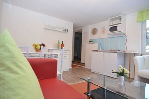 Una sala de estar moderna con un sofá rojo, una mesa de centro de vidrio y una zona de cocina con armarios blancos y un microondas.