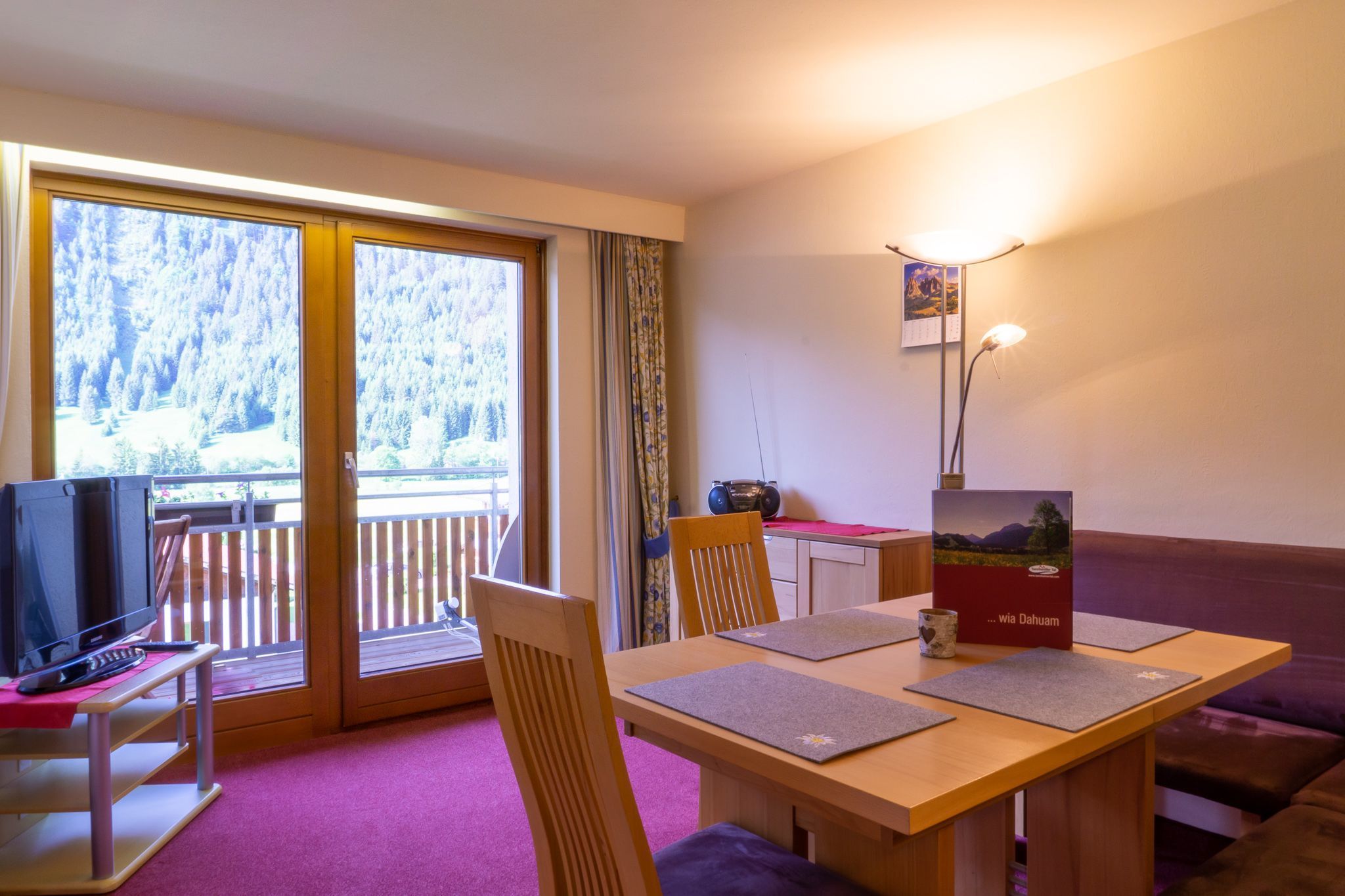 Quarto com mesa de madeira, cadeiras, televisão, sofá, varanda com vista para árvores e uma luminária.