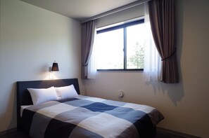 Un lit bien fait, recouvert d’une couvre-lit à carreaux, une lampe de chevet et une fenêtre avec des rideaux.