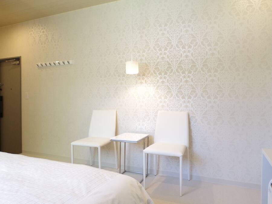 Une chambre d’hôtel avec un lit, deux chaises blanches, une petite table et un mur décoré d’un motif.