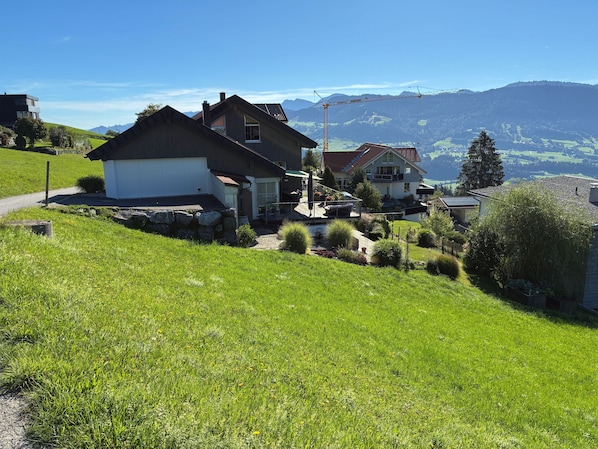 A residential area with two houses on a hillside, surrounded by greenery and a clear view of distant mountains.