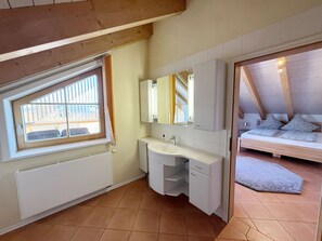 Ein Schlafzimmer mit Bett, Fenster, Badezimmer mit Waschbecken und Spiegel sowie Blick ins Freie.