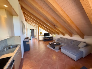 Ein modernes Loft mit Küche, Wohnbereich inklusive Sofa und Fernseher sowie einer hohen Decke mit freiliegenden Balken.