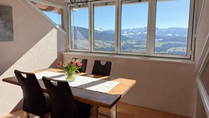 Un espace repas comprenant une table, des chaises et une vue sur les montagnes, visible à travers une grande fenêtre.