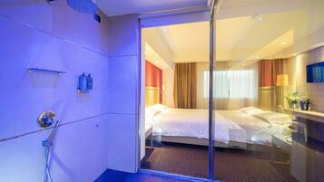 Een hotelkamer met een douchecabine van glas, een bed met witte lakens, een nachtkastje met een lamp en een aan de muur bevestigde televisie.