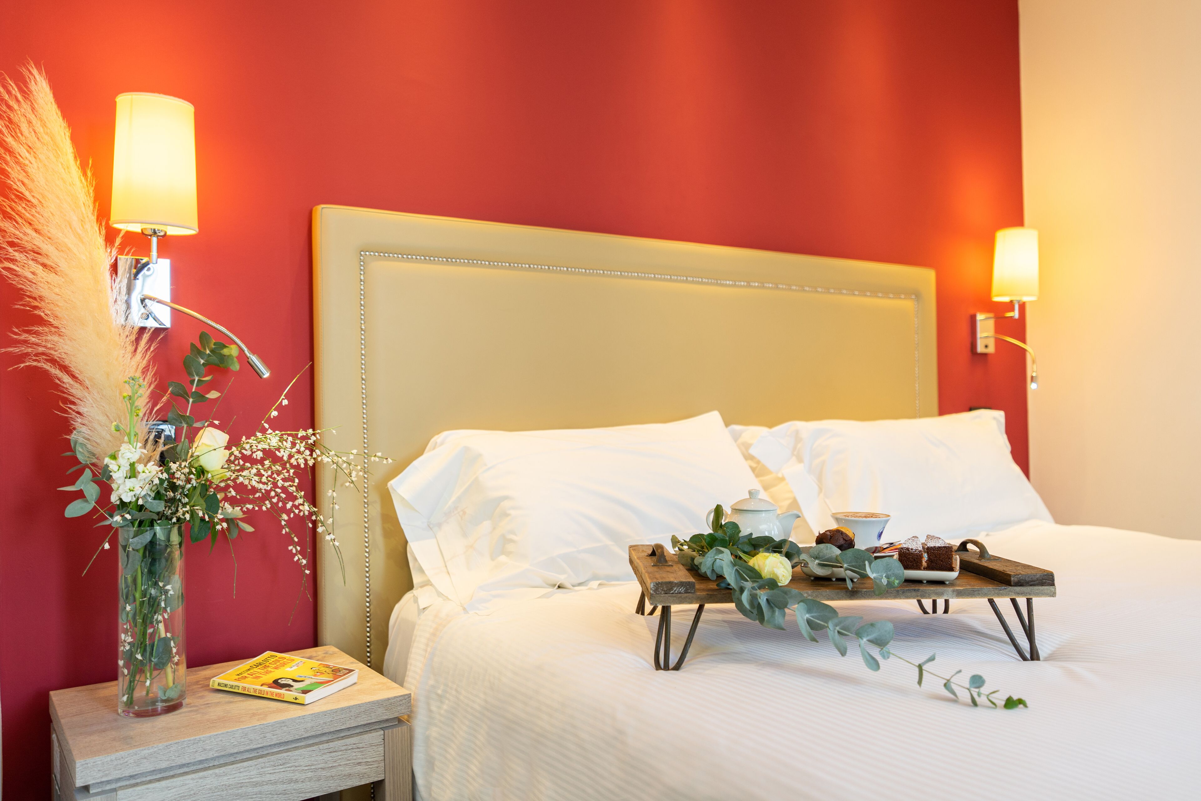 A neatly made bed with white linens, a bedside table with a book and a vase of flowers, and a tray with a teapot and pastries.