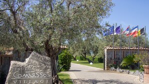Une pancarte en pierre portant le nom « Tenuta Comisso Village » et un chemin pavé menant à un bâtiment où flottent plusieurs drapeaux.