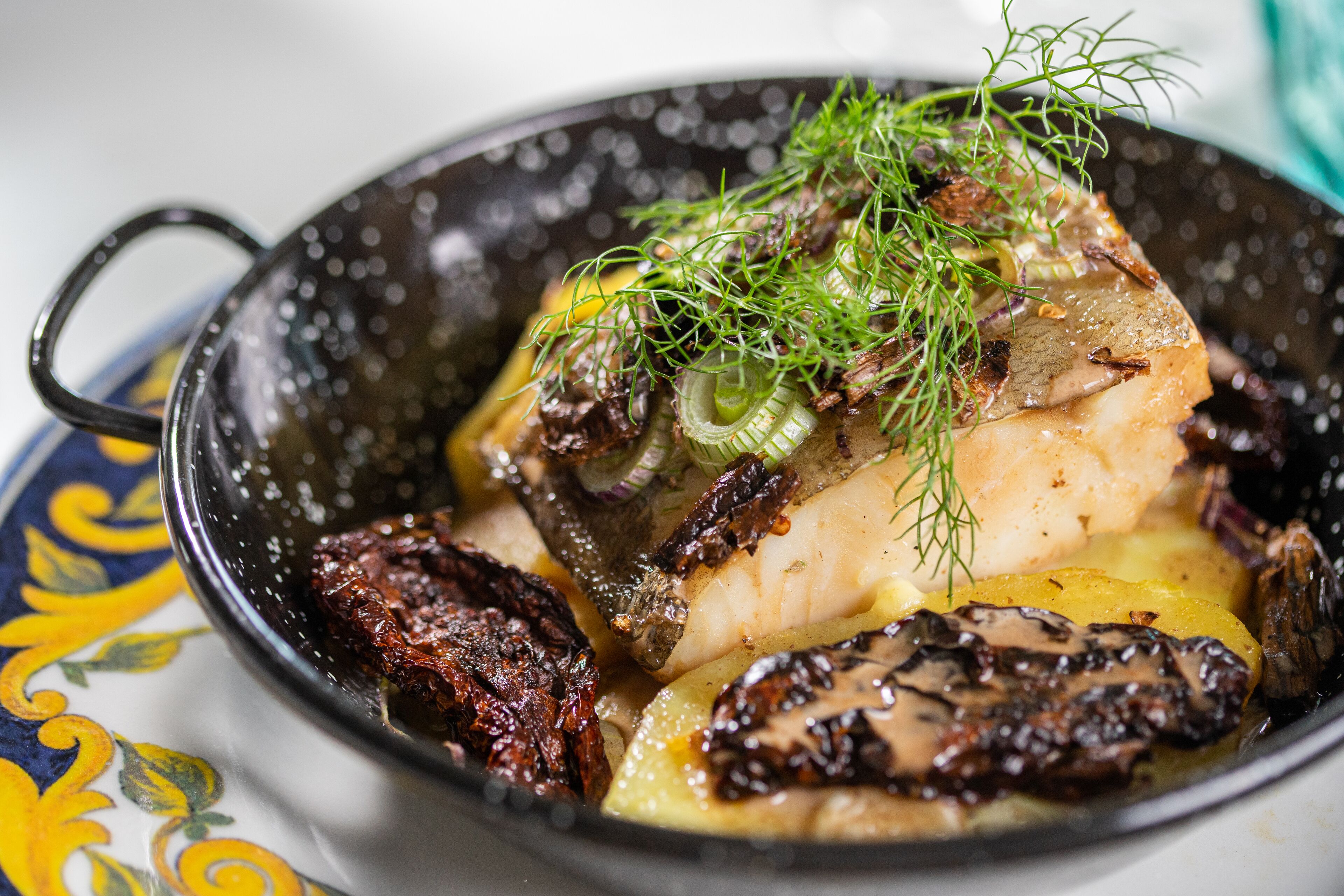 A bowl of food with a handle, containing sliced fish, mushrooms, and garnished with herbs.