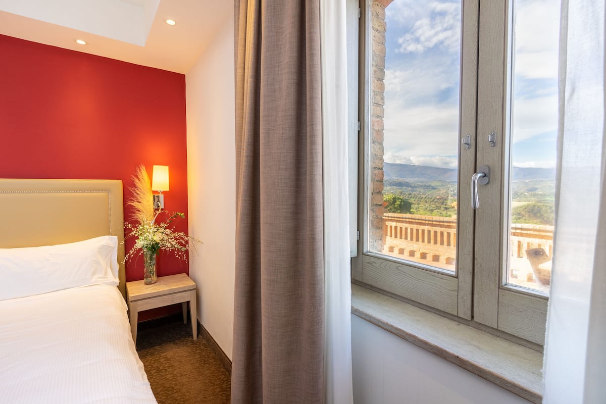 Une chambre d’hôtel avec un lit, une table de chevet avec une lampe et des fleurs, et une fenêtre donnant sur un bâtiment et un paysage.