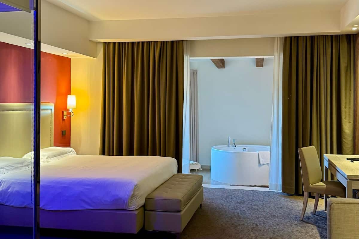 Une chambre d’hôtel comprenant un lit, un bureau, une chaise, une salle de bain avec baignoire et une grande fenêtre avec des rideaux.
