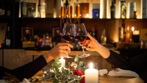 Une scène de bar avec deux personnes qui trinquent avec des verres de vin, une bougie et une table recouverte d’une nappe blanche, ornée d’une rose rouge.