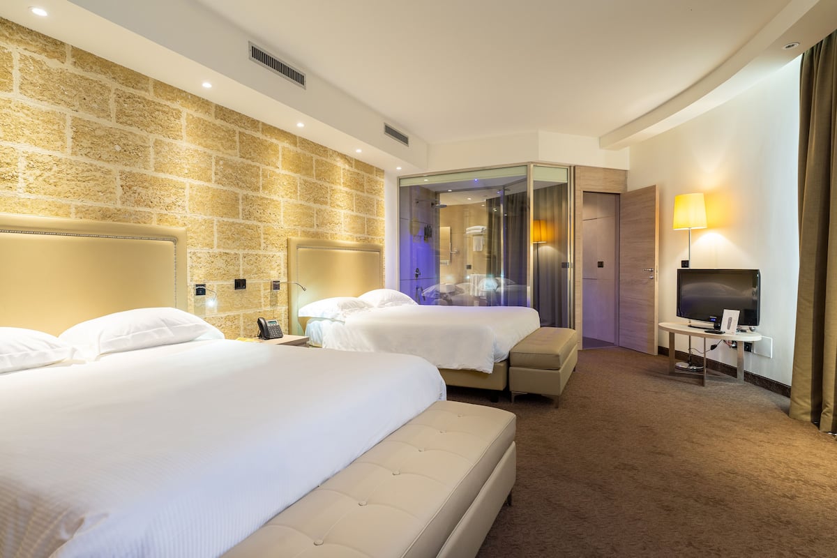 Une chambre d’hôtel avec deux lits, une télévision, un bureau et une salle de bain séparée par une paroi en verre.