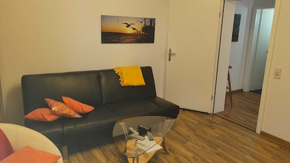 A living room with a black leather sofa, two orange pillows, a glass coffee table with remotes and a magazine, and a painting of birds flying over water on the wall.