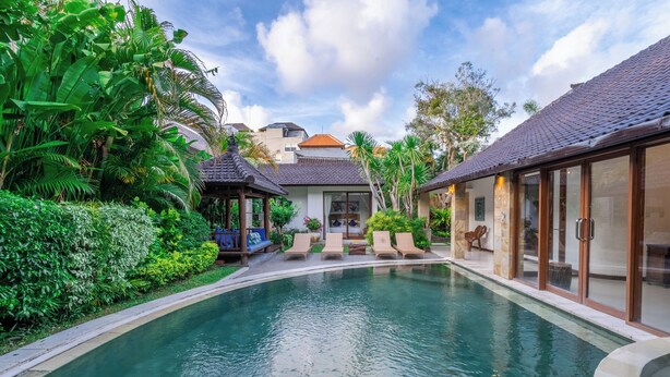 A swimming pool with lounge chairs, surrounded by lush greenery and a building with a tiled roof. - Villa Aveli Seminyak by Best Deals Asia Hospitality (Seminyak)