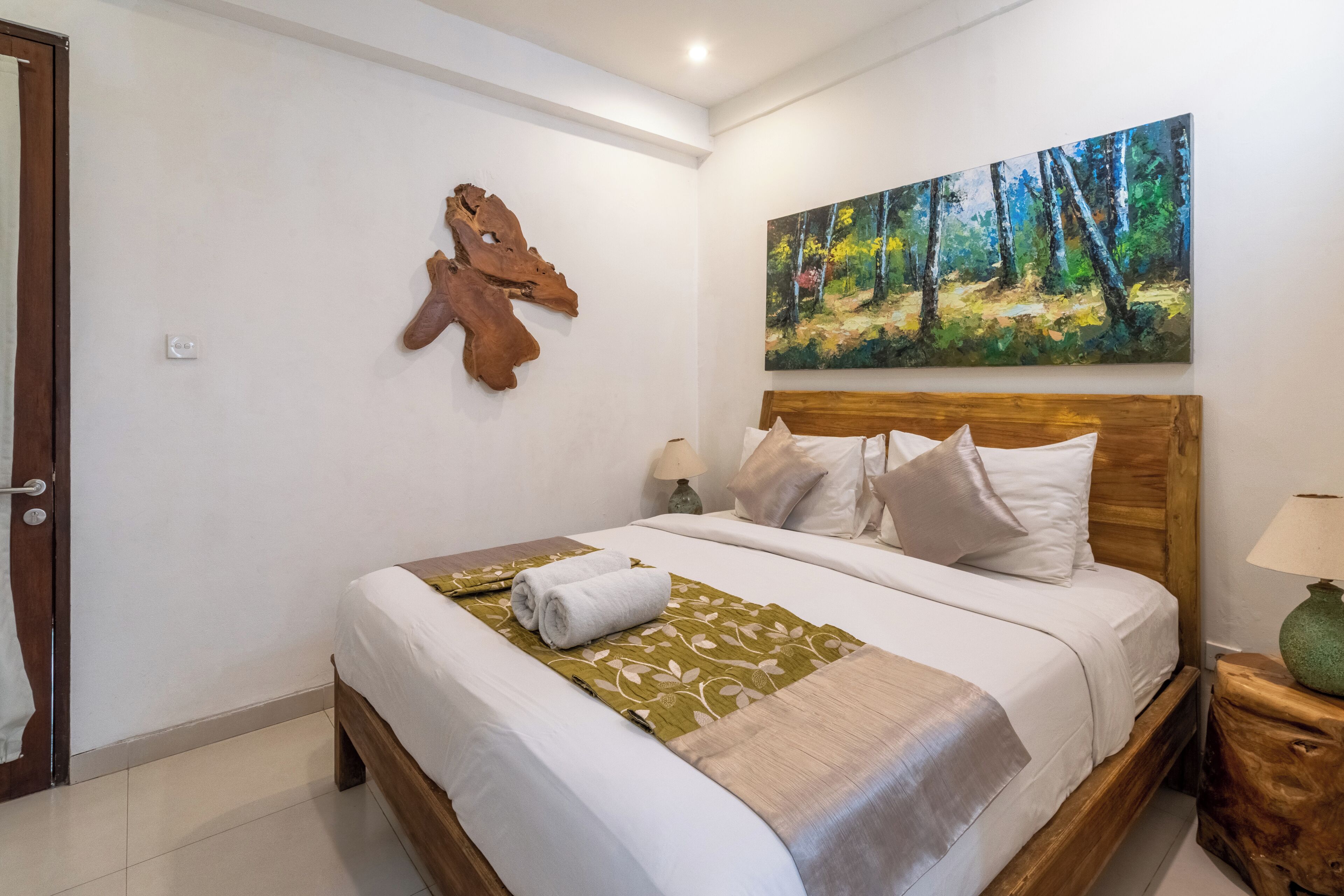 Un lit bien fait, agrémenté d’un couvre-lit à motifs floraux verts, d’une tête de lit en bois et d’un tableau représentant une forêt accroché au mur.