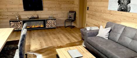 Una sala de estar acogedora con chimenea, un sofá gris, una mesa de centro de madera y un televisor montado en la pared.