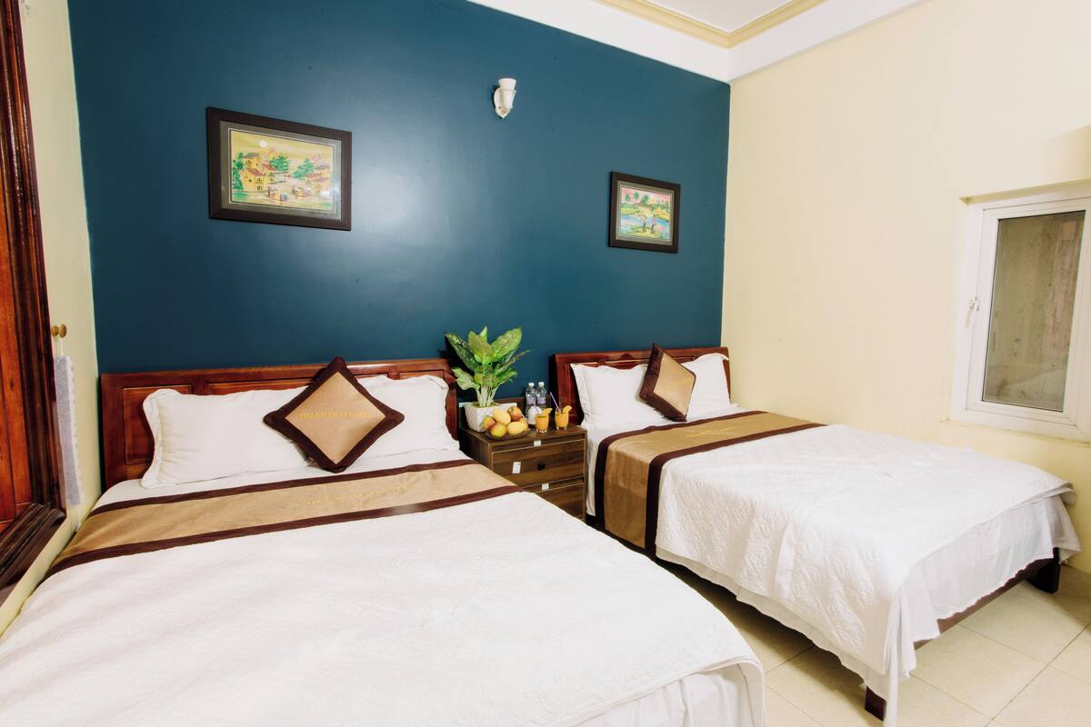 Een hotelkamer met twee bedden, een nachtkastje met een plant en fruit, en twee foto’s in lijsten aan de muur.