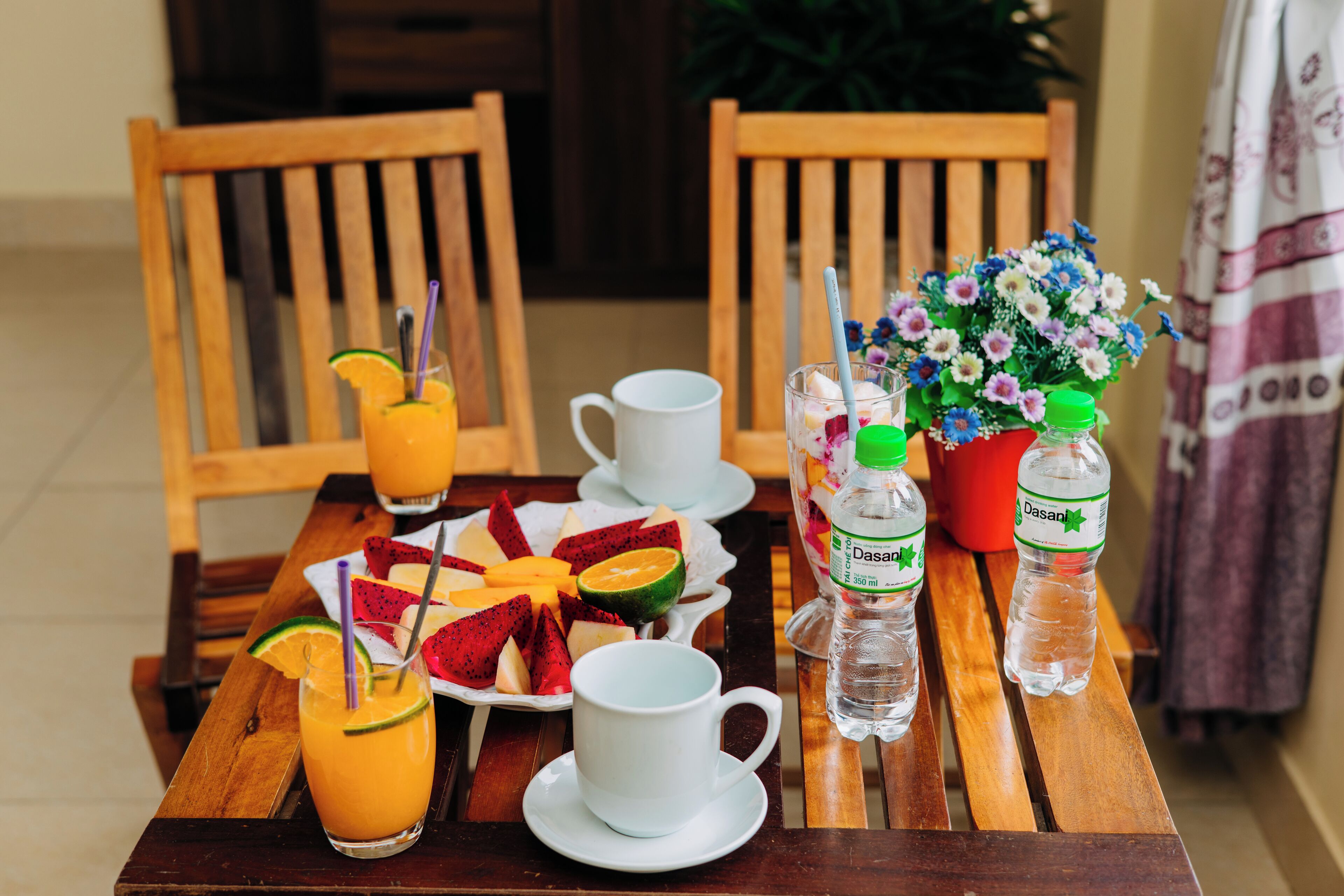 Una mesa de madera con una bandeja de frutas, dos tazas blancas, un vaso de jugo de naranja y dos botellas de agua Dasani.