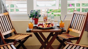 Un coin repas avec des chaises en bois et une table dressée pour deux personnes, une fenêtre avec des rideaux et une vue sur la verdure et un bâtiment au loin.