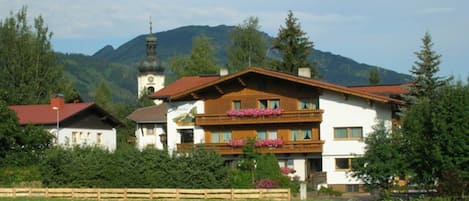 A multi-story building with balconies, surrounded by greenery and mountains.