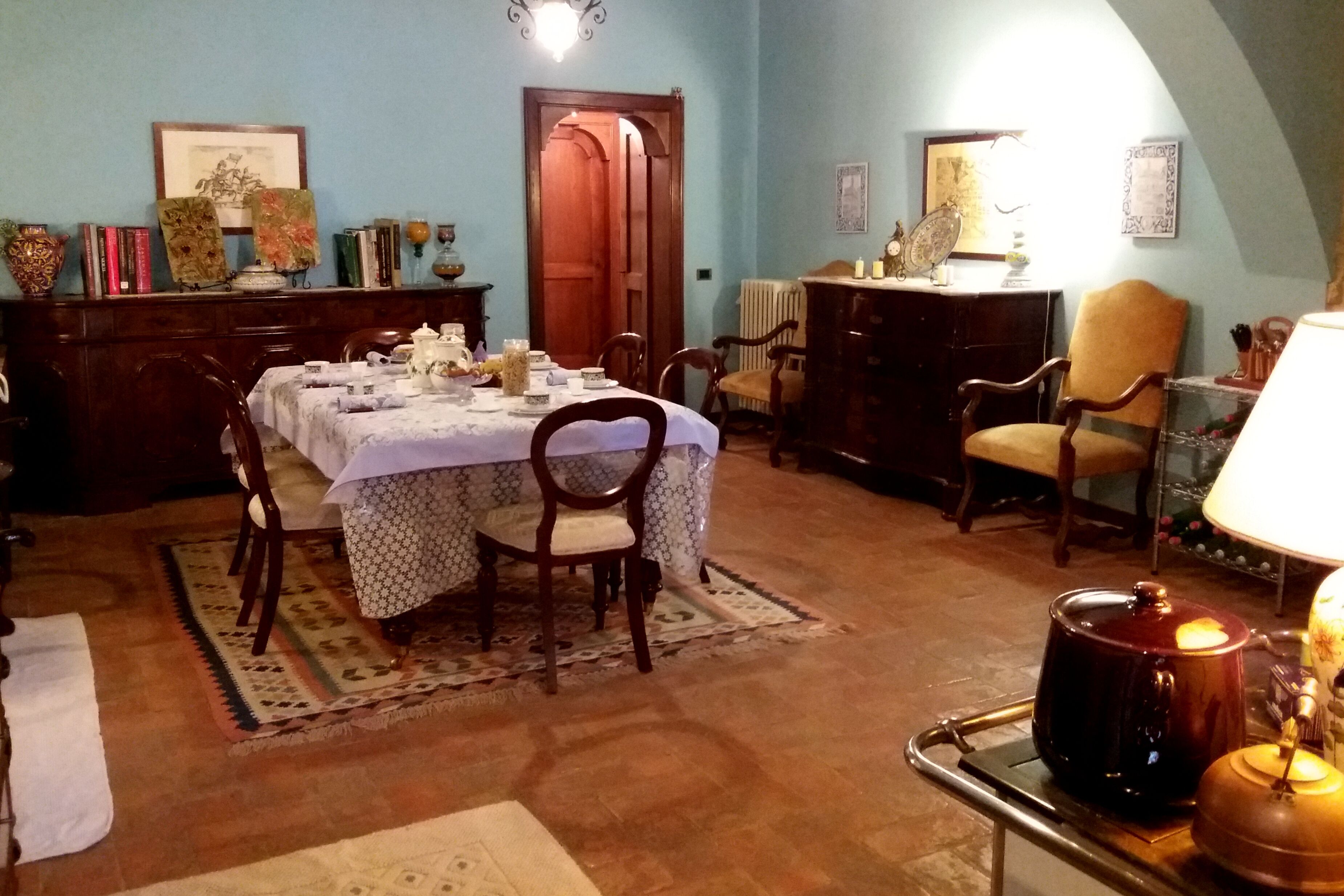 A dining area with a table set for four, surrounded by wooden chairs. A radiator is visible in the background, and a wooden cabinet with decorative items is to the left.
