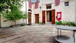 Un patio con suelo de ladrillo, una mesa redonda y un edificio con puertas y contraventanas de madera.
