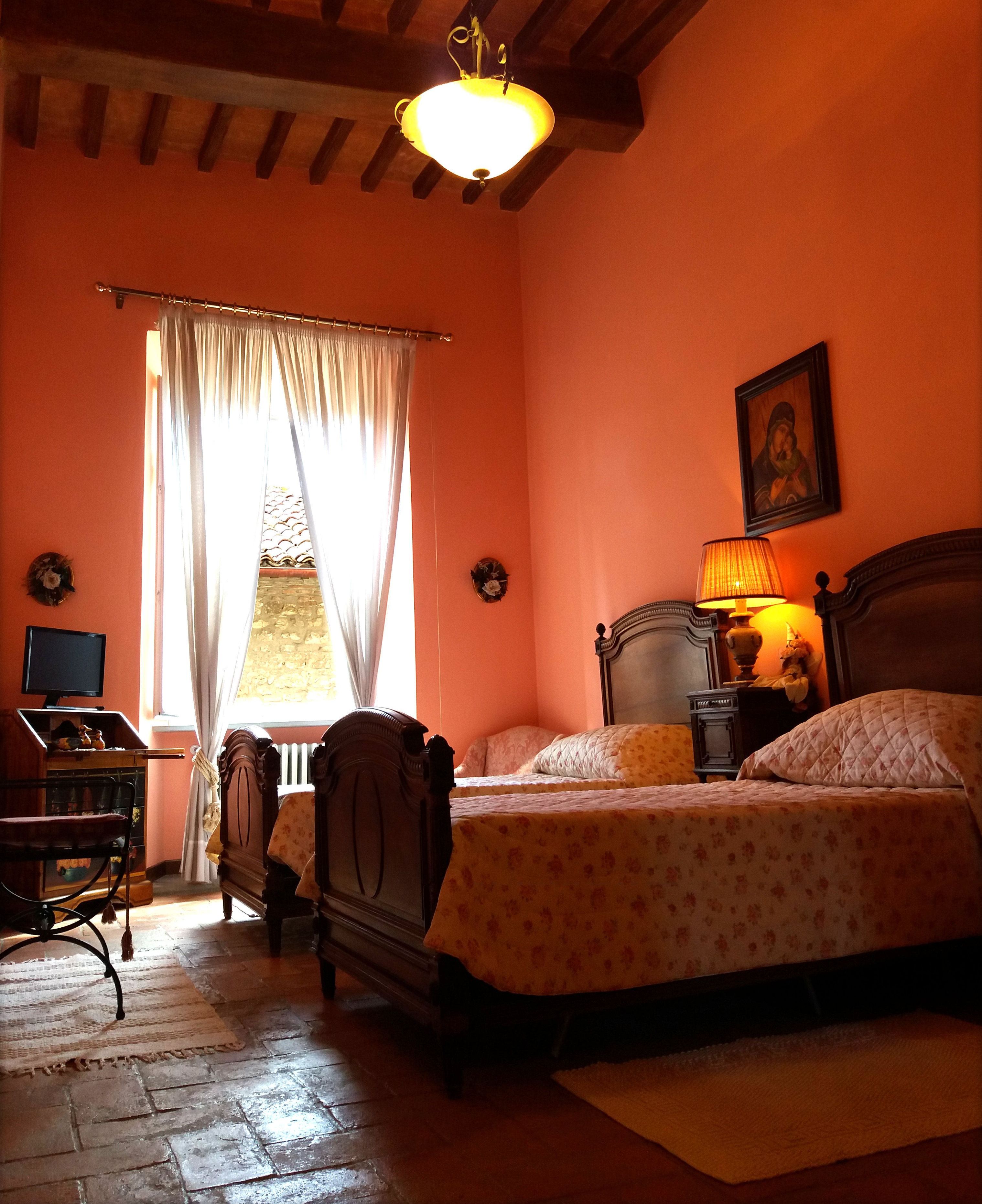 A bedroom with a wooden bed, a nightstand with a lamp, a chair, a desk, and a window with curtains.