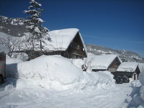 Una cabaña de madera cubierta de nieve con techo empinado, rodeada de aluviones y un cielo azul despejado.