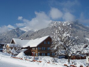 Un edificio de dos pisos con tejado cubierto de nieve, rodeado de árboles nevados y montañas al fondo.