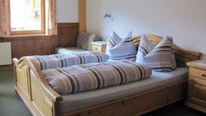 Una cama doble con ropa de cama a rayas, un cabecero de madera, una mesita de noche y una ventana con cortinas. Se observa una cruz en la pared.
