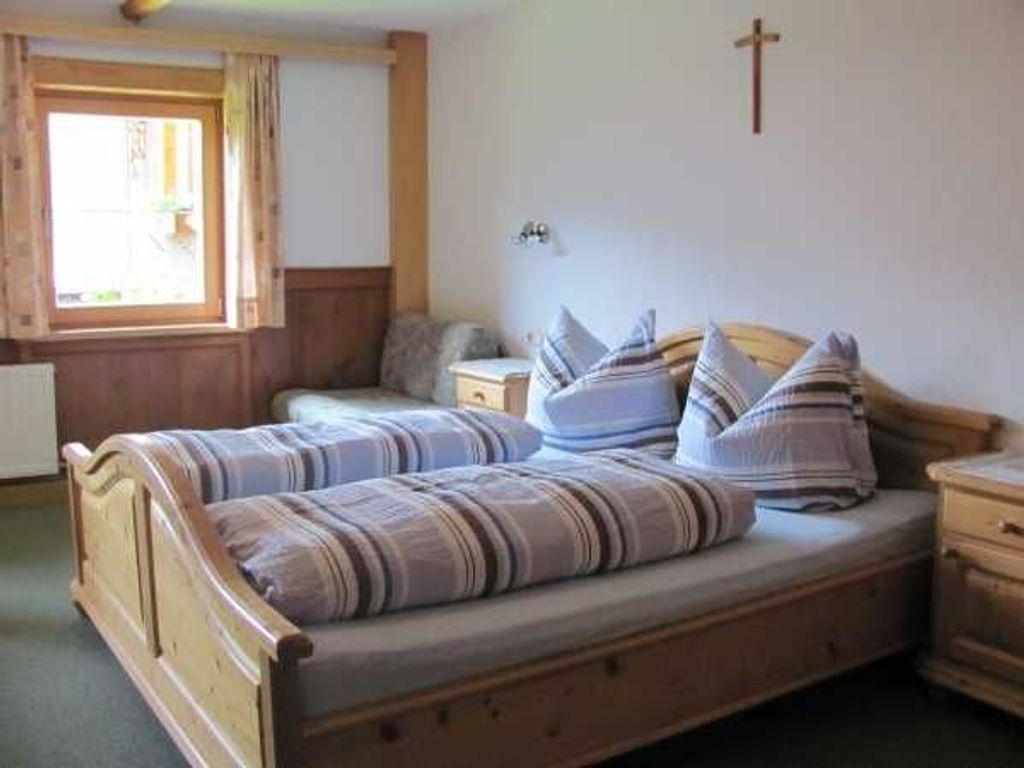 Una cama doble con ropa de cama a rayas, un cabecero de madera, una mesita de noche y una ventana con cortinas. Se observa una cruz en la pared.