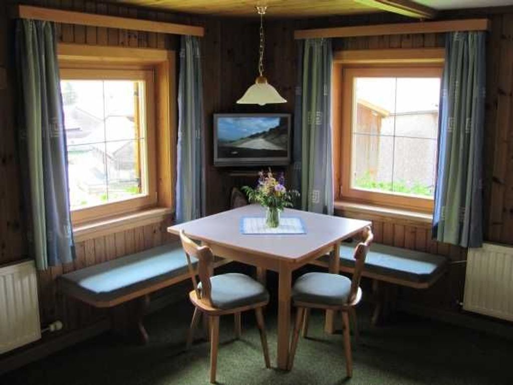 A room with a wooden table, four chairs, a bench, a television, and windows with curtains.