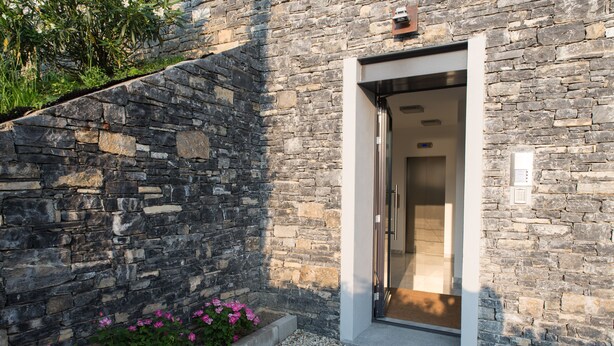 Un edificio de piedra con entrada de vidrio, un camino de grava y flores rosadas.