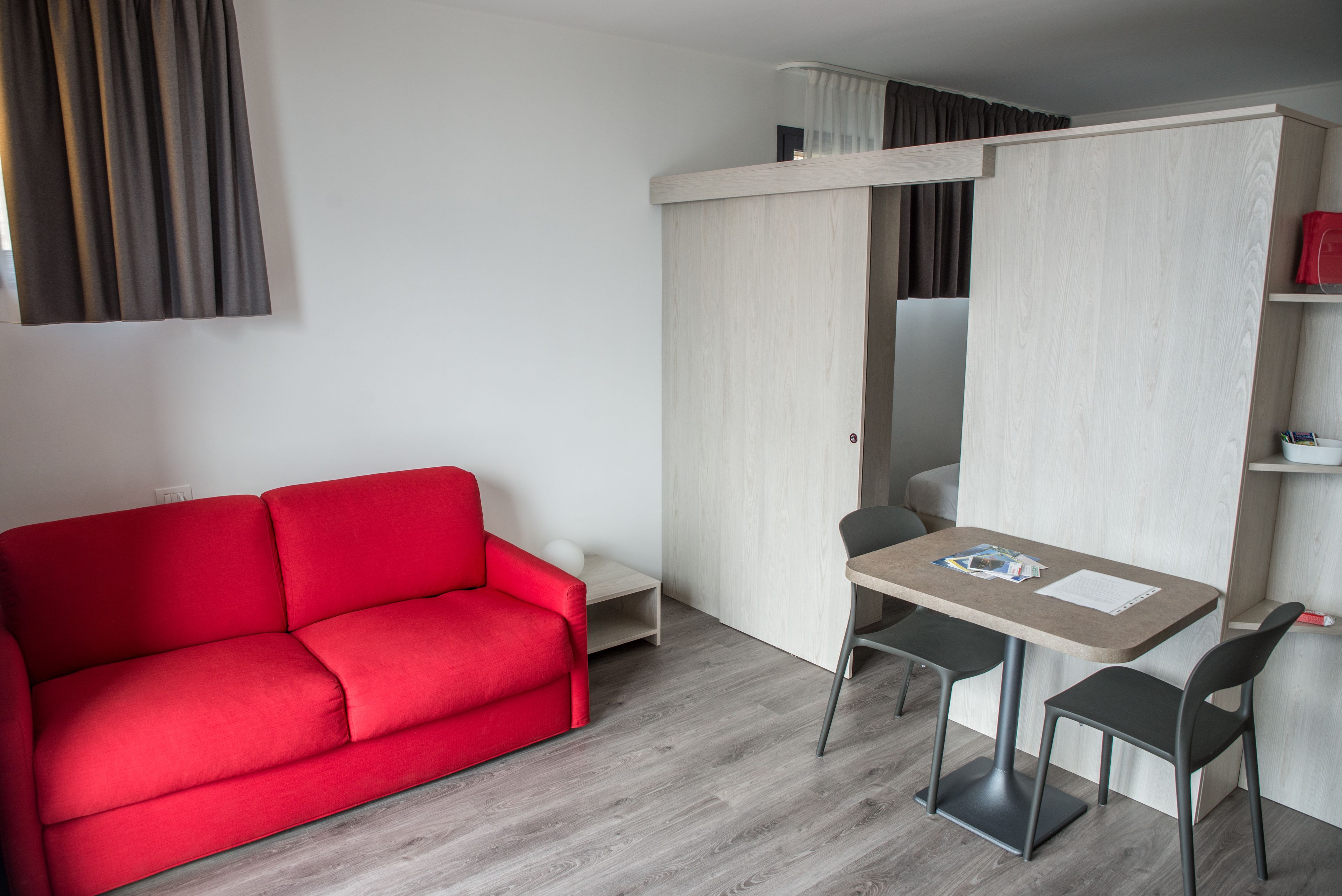 Une pièce de taille modeste, avec un canapé rouge, une petite table accompagnée de deux chaises et une armoire à porte coulissante.