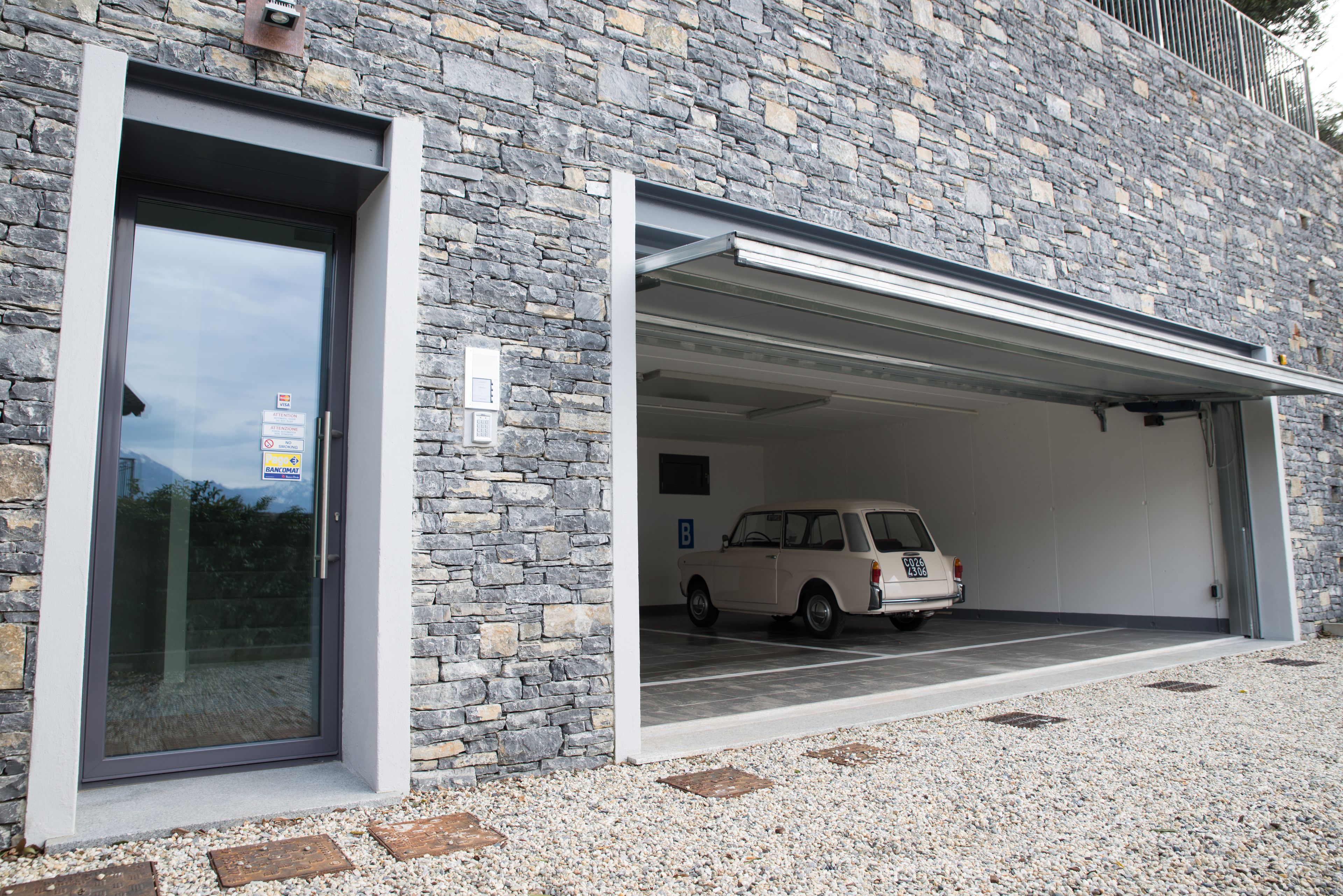 Ein modernes Gebäude mit einer Stein-Fassade, eine Garage mit einem weißen Auto darin und eine Schotterzufahrt.