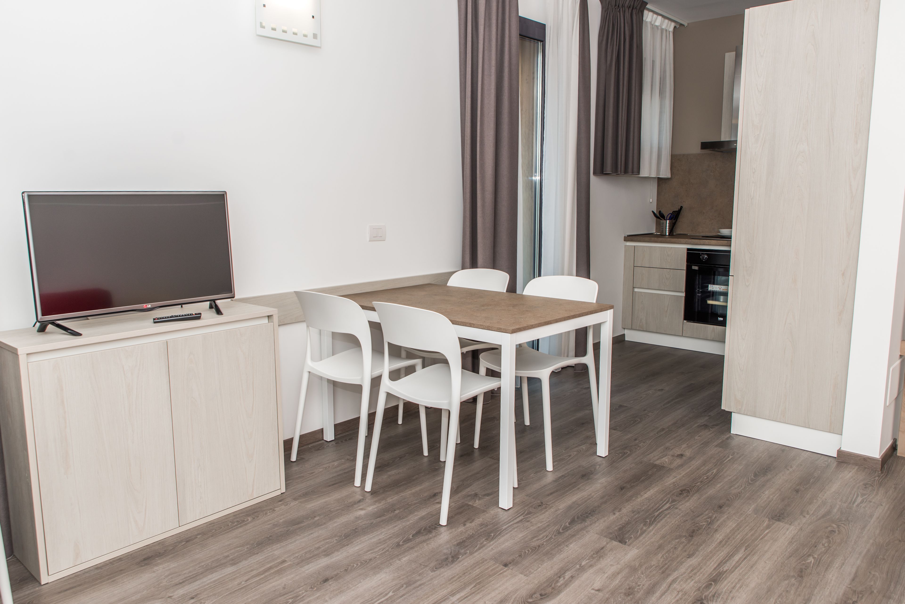 A modern kitchen with a dishwasher, a dining area with a wooden table and white chairs, and a TV on a cabinet.