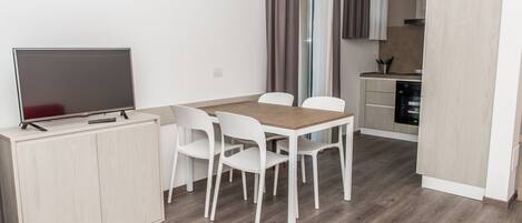 A modern kitchen with a dishwasher, a dining area with a wooden table and white chairs, and a TV on a cabinet.