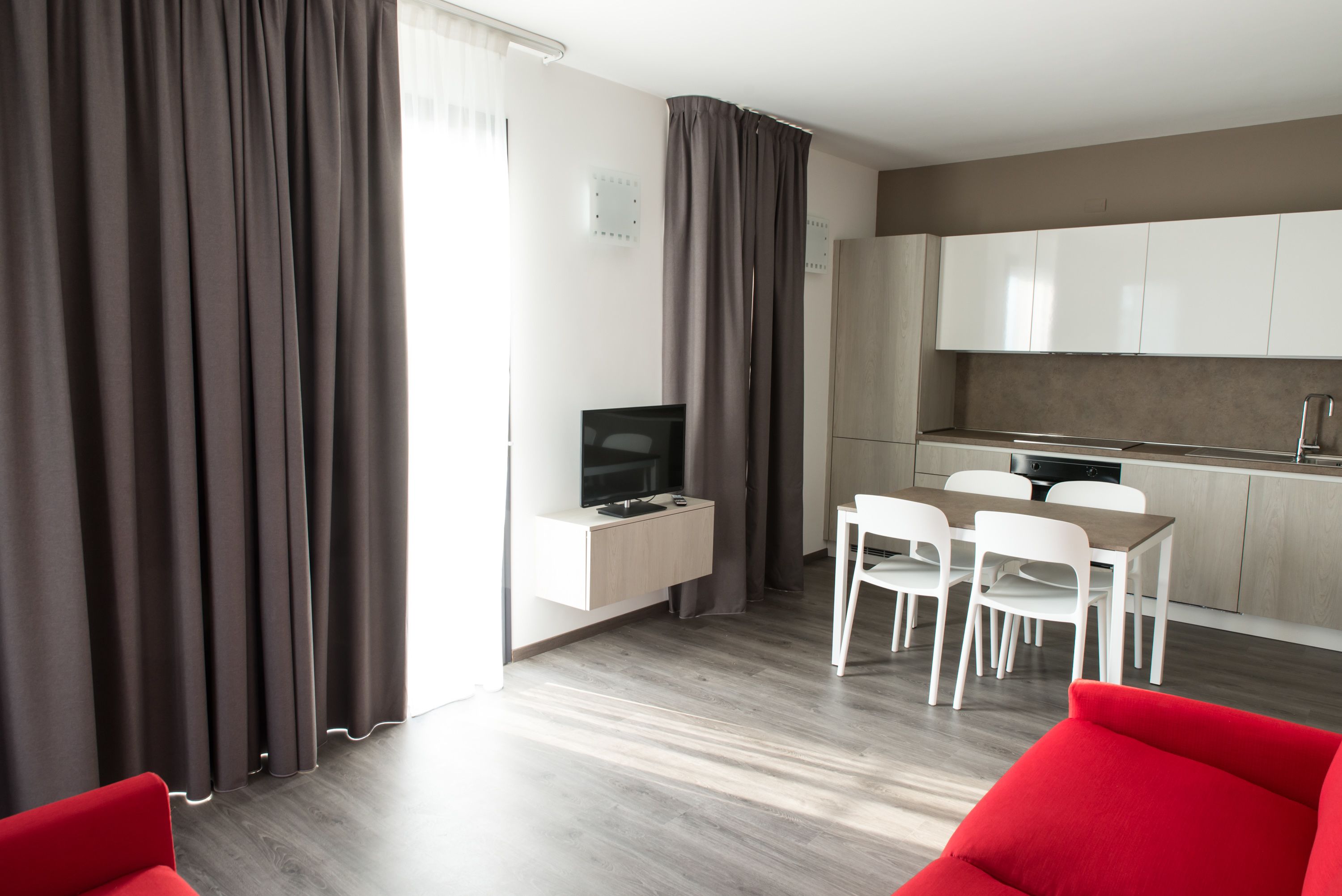 A modern living room with a flat-screen TV, a dining area with white chairs, and a kitchen with wooden cabinets.