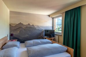 Une chambre d’hôtel avec une fresque représentant une montagne, un lit avec une literie à carreaux, un téléviseur posé sur un meuble en bois et une fenêtre avec des rideaux.
